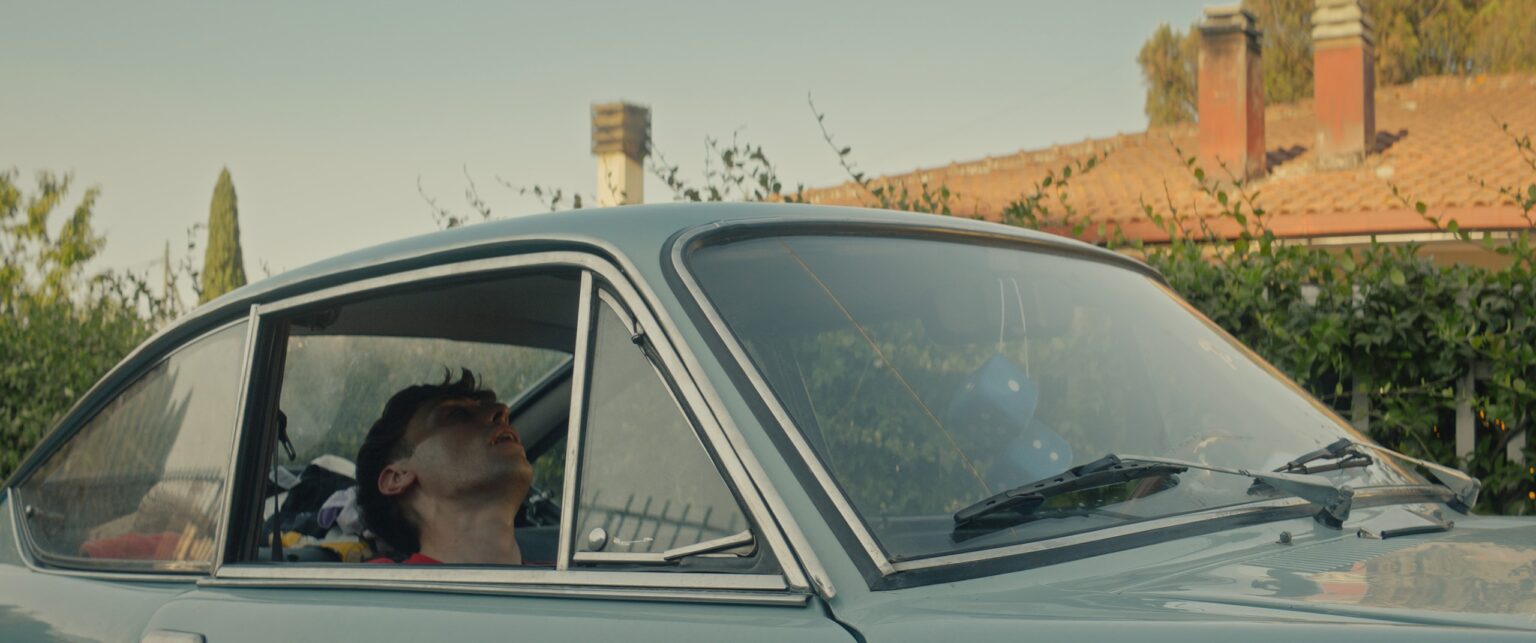La Truffa, directed by Matteo Stipa and Michele Marchionne: a young man with his head tilted back inside a pale blue vintage car, in front of a hedge and a tiled roof