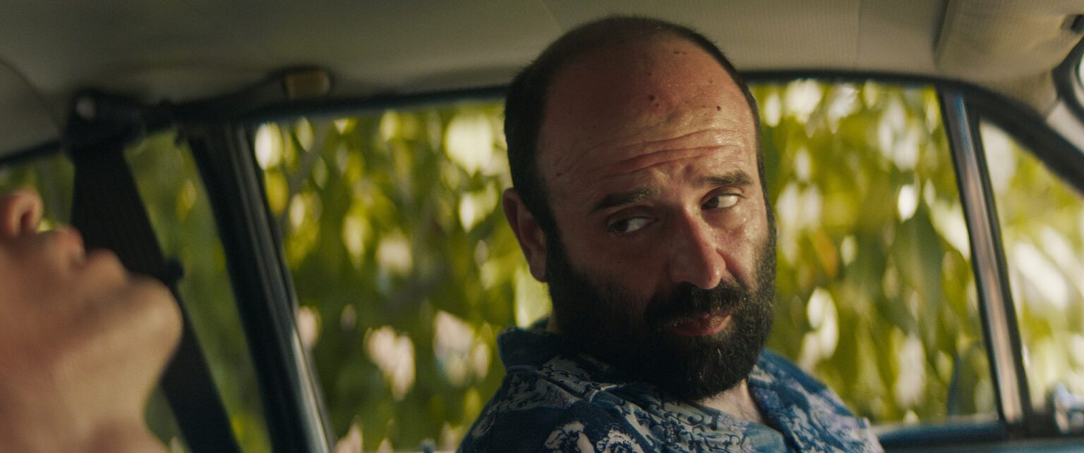 La Truffa, directed by Matteo Stipa and Michele Marchionne: Frappa, bearded, in a floral shirt, looks sideways in the car