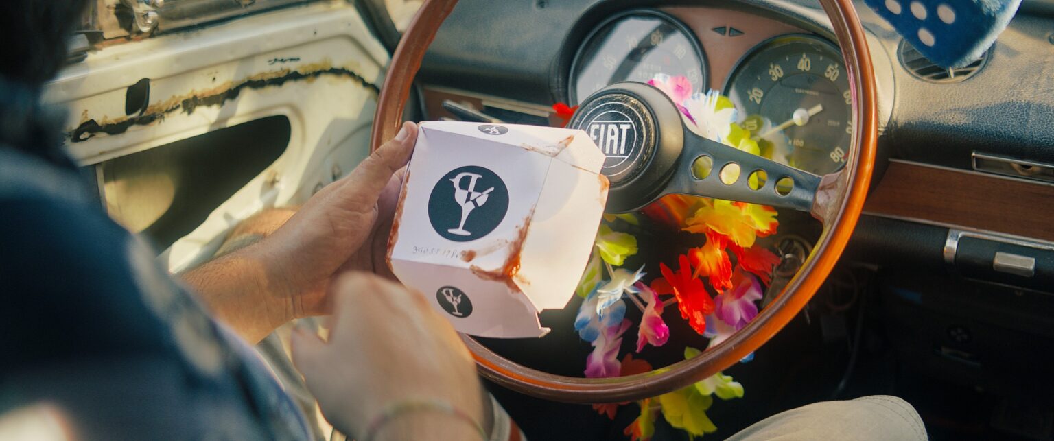 La Truffa, directed by Matteo Stipa and Michele Marchionne: a takeaway box in front of the steering wheel, with a flower garland hanging