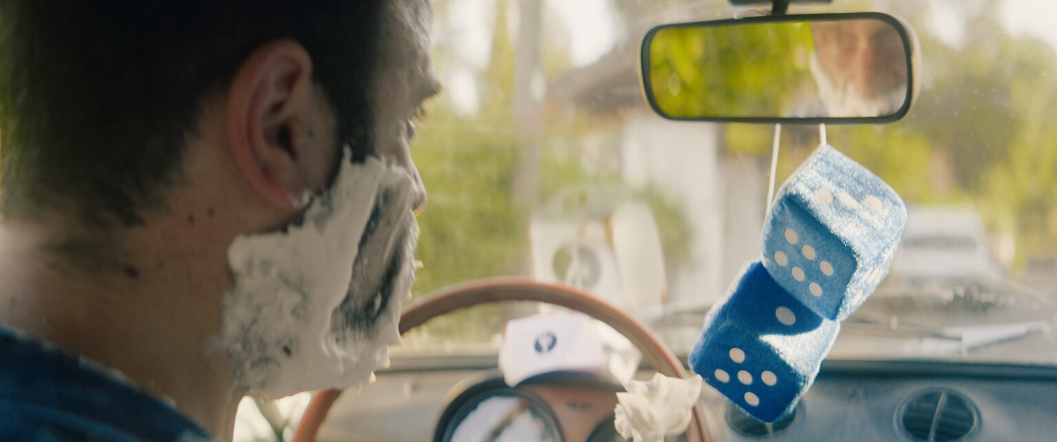 La Truffa, directed by Matteo Stipa and Michele Marchionne: a man with shaving foam on his face, in the car, with fluffy blue dice hanging from the mirror
