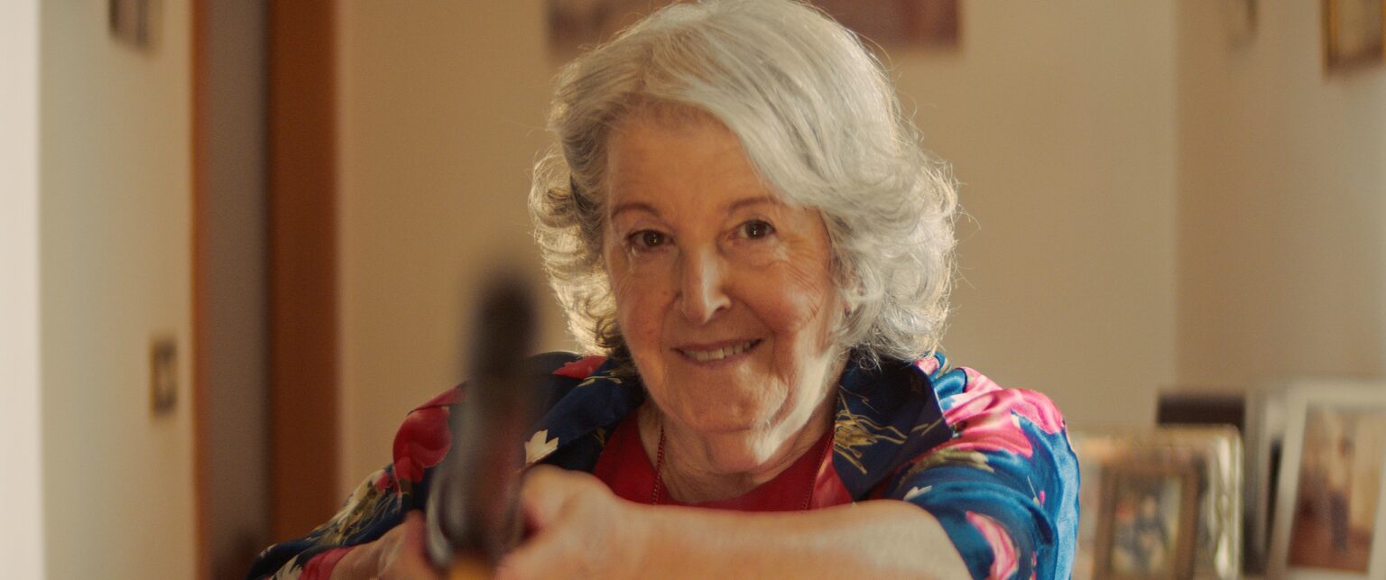 La Truffa, directed by Matteo Stipa and Michele Marchionne: Mirella smiles in the living room, with a blurred rifle in the foreground